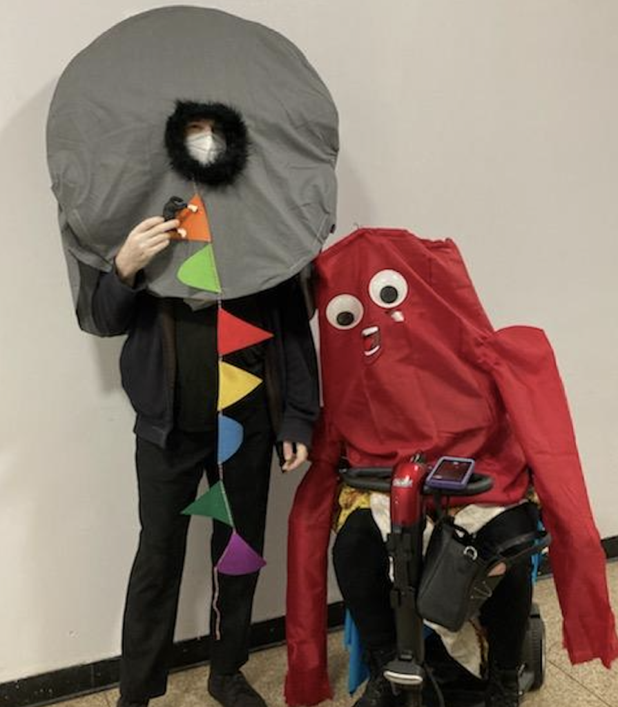 My husband and I posing in our Halloween Nope costumes. My husband is the creature, he's wearing a large grey circular object on his head with black fur trim around where his head sticks out in the middle. He's holding a small plastic horse and has party flags hanging from the hole. I'm sitting in my scooter, you can't really see me besides my legs in black pants. I'm dressed as one of the blow-up, dancing car dealer figures. The costume is all red with extra large arms, googly eyes, and a wide open mouth sticker.