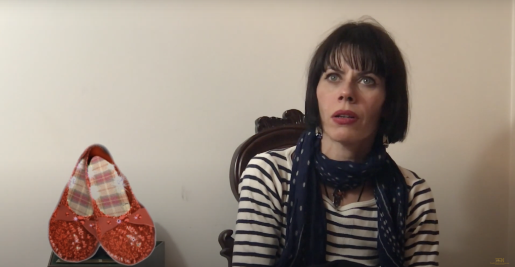 A screenshot from documentary Remembering Return to Oz features actor Fairuza Balk with a pair of ruby slippers to her left. She's got black hair in a bob style with bangs and is wearing a black and white horizontally striped shirt with a navy blue scarf.