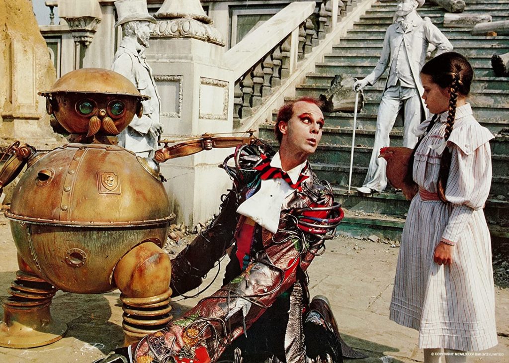 An image from the film Return to Oz. Round, bronze mechanical soldier Tik-Tok holds the Head Wheeler captive while Dorothy interrogates him. The Wheeler is wearing a red and black striped bowtie and has elongated arms, Dorothy is holding her chicken Belina and is has her hair styled in  pigtails.