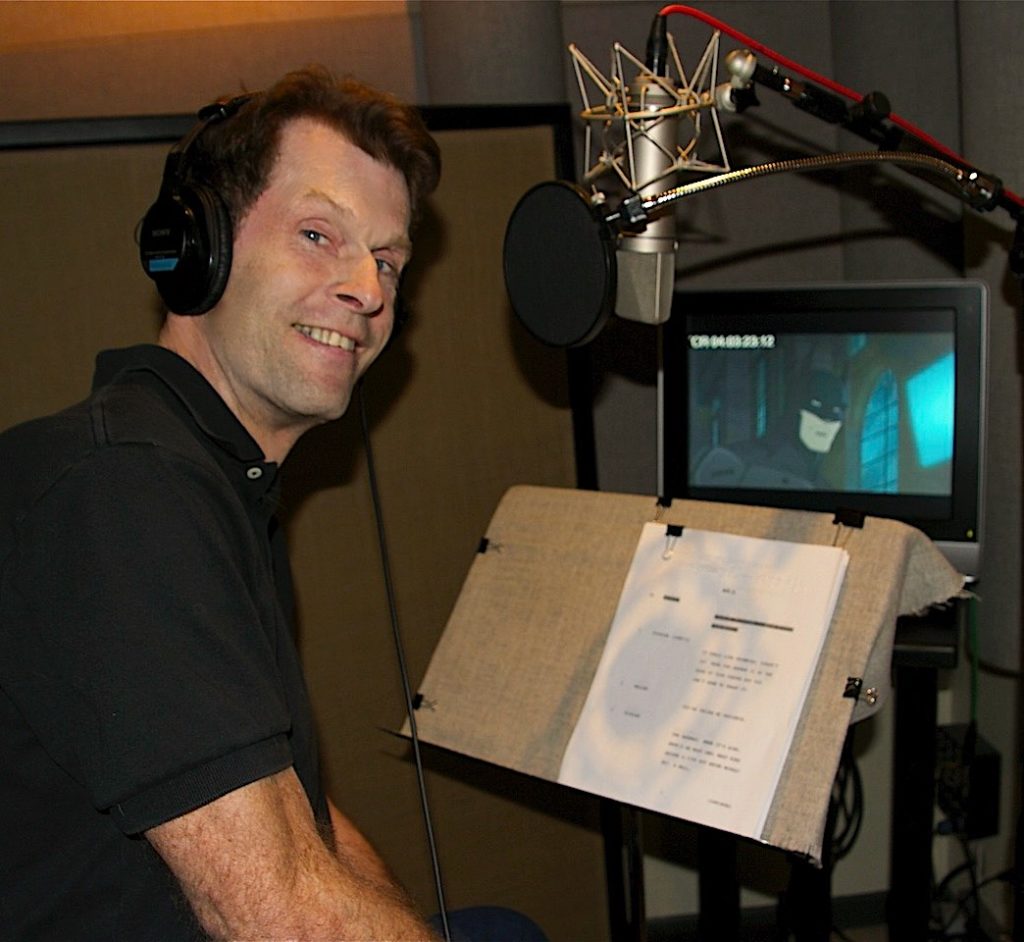Batman voice actor Kevin Conway looks at the camera while sitting in a recording booth. He's wearing headphones and has his script, a microphone, and a still of Batman from an animated film on a screen in front of him.