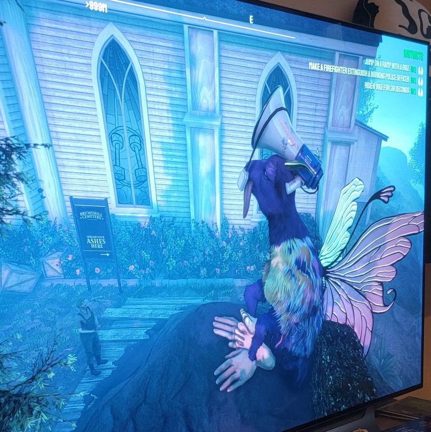 A photo I took of my TV while playing Goat Simulator 3. My goat was giant-sized at the time and is purple with green, blue, and purple on the chest area. Its wearing a bullhorn on its head and blue and pink fairy wings on its back. It's sitting next to a church and on a pile of ashes.