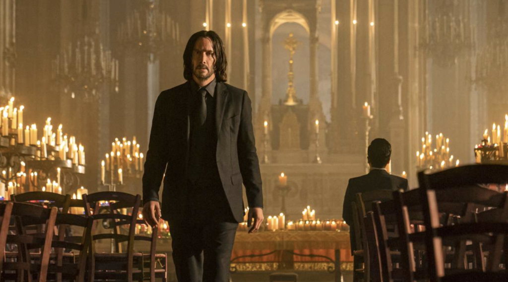 Actor Keanu Reeves is walking away from a church altar lit by a ridiculous number of candles. He's wearing his trademark all-black suit. There is a man sitting in a chair facing away from him with dark hair and a dark suit.