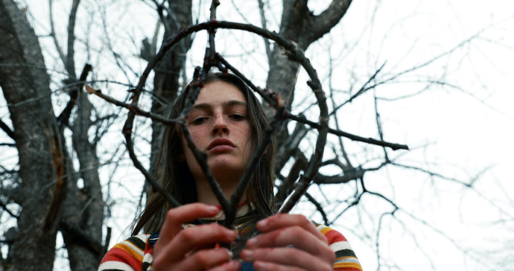 Actor Zelda Adams from the film Hellbender. It's a shot from below in the woods and we see her face through a witchy creation of twigs.